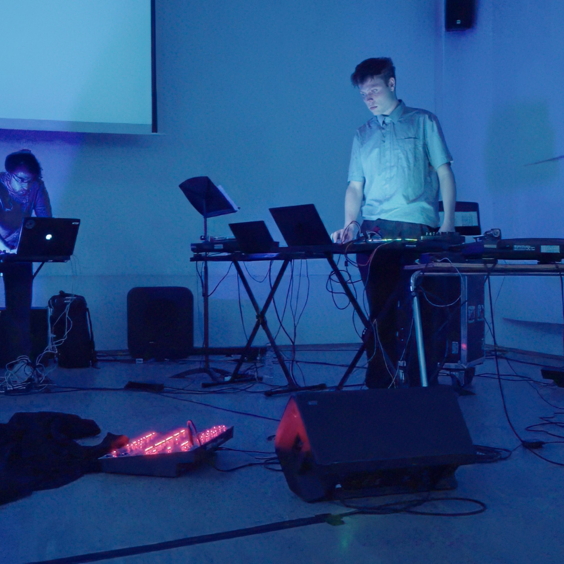 Zwei Männer stehen in einem Studio an Synthesizern. Es ist dunkel und es leuchten Laptops und ein Mischpult.