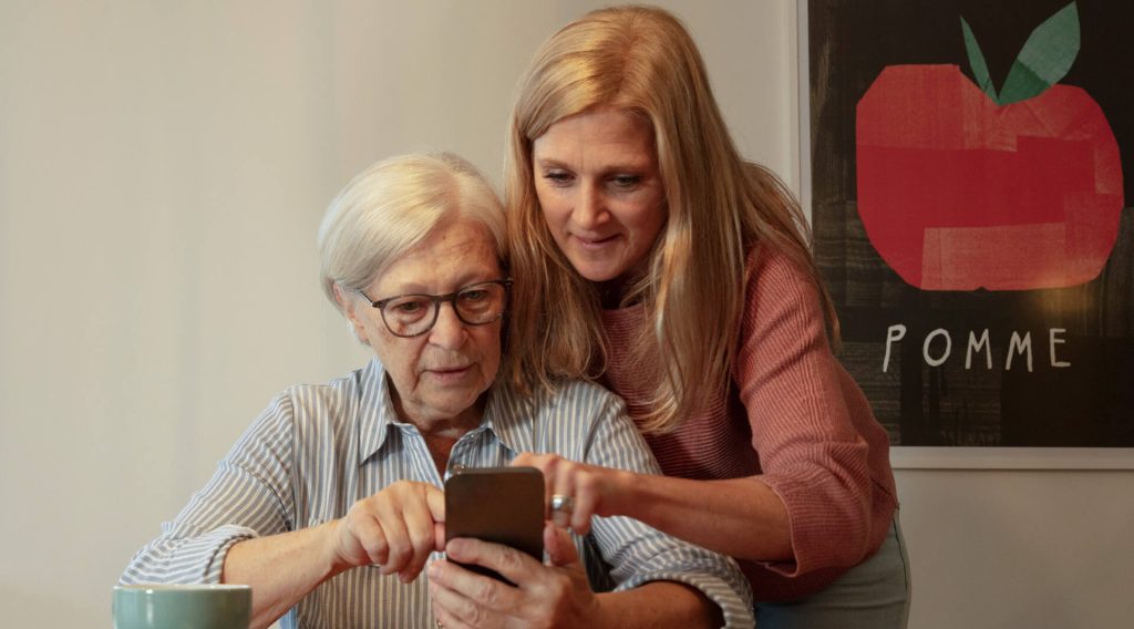 Eine jüngere Frau hilft einer älteren Frau bei der Bedienung des Smartphones.