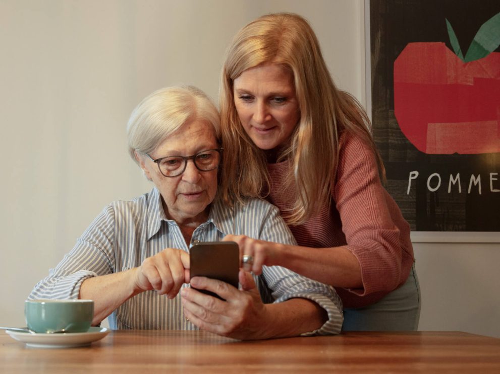 Eine jüngere Frau hilft einer älteren Frau bei der Bedienung des Smartphones.