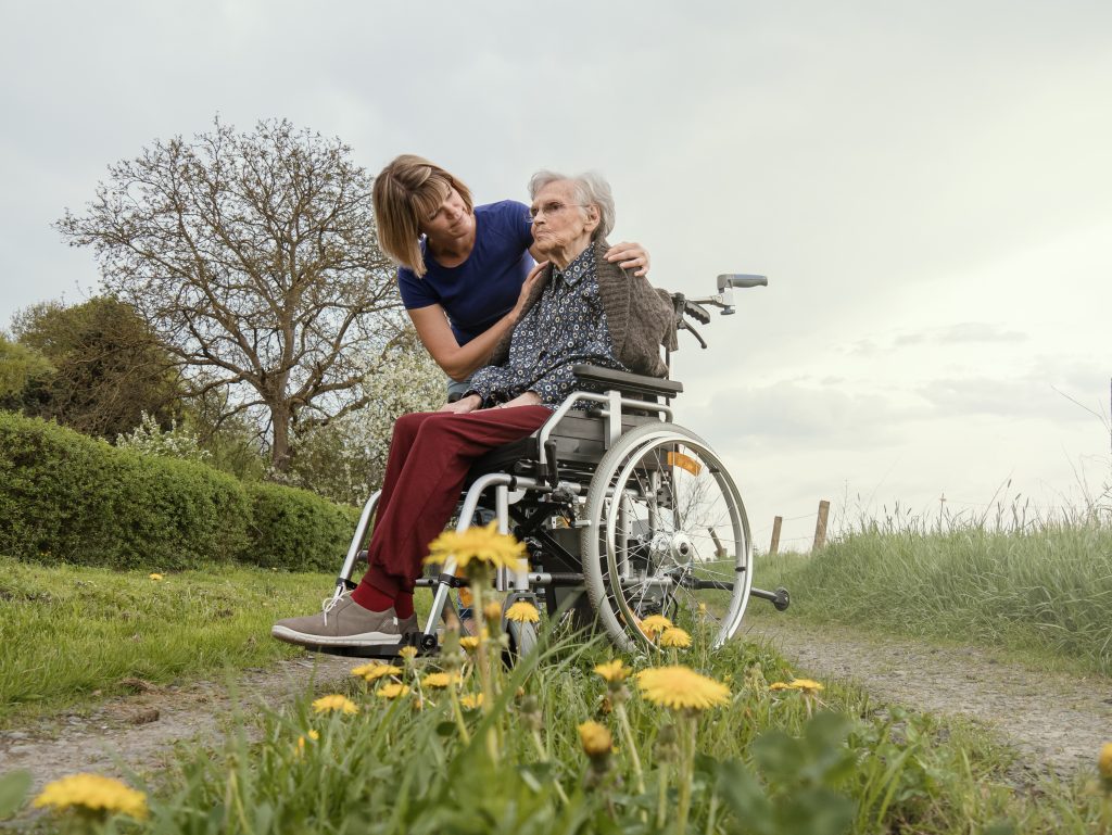 Eine ältere Person sitzt im Rollstuhl auf einem Weg in der Natur und wird von einer Frau betreut.