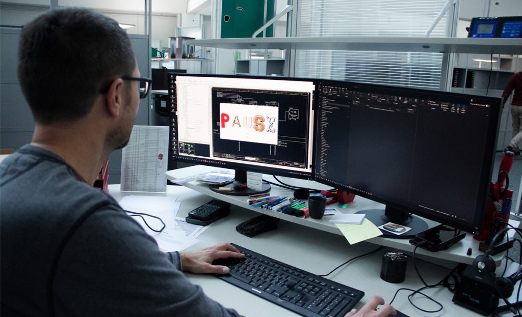 Mitarbeitender am Schreibtisch, zwei Monitore; auf einem erscheint gross «Pause» als Desktop-Erinnerung.