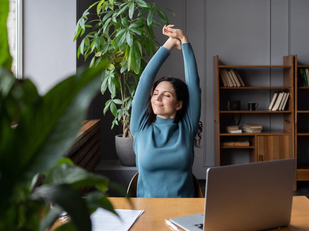 Frau, am Schreibtisch vor dem Laptop sitzend, streckt sich genüsslich