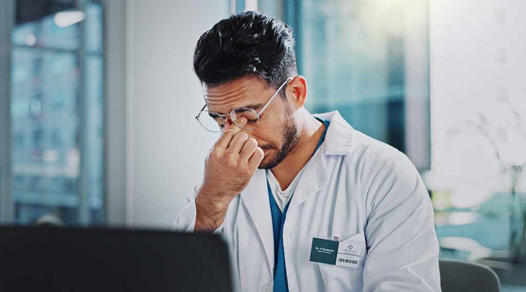 Übermüdeter Arzt in Spitalkleidung am Computer beim Schreiben von Krankenberichten.