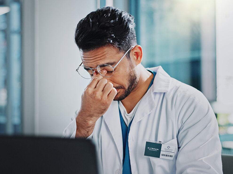 Übermüdeter Arzt in Spitalkleidung am Computer beim Schreiben von Krankenberichten.