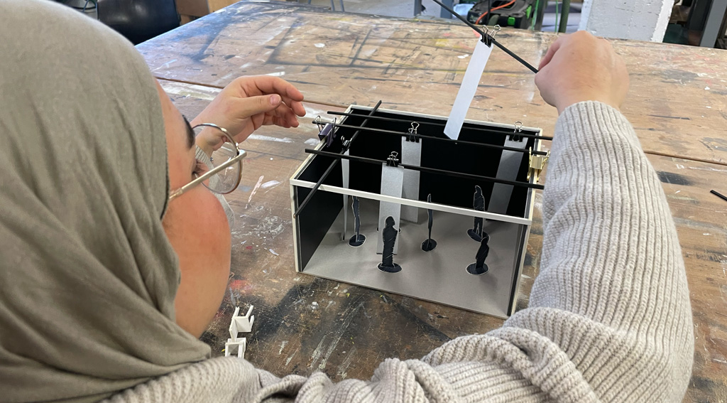 Eine Person arbeitet an einem Arbeitsmodell mit mehreren transparenten Flächen und kleinen Figuren auf einem Tisch in einem Atelier.