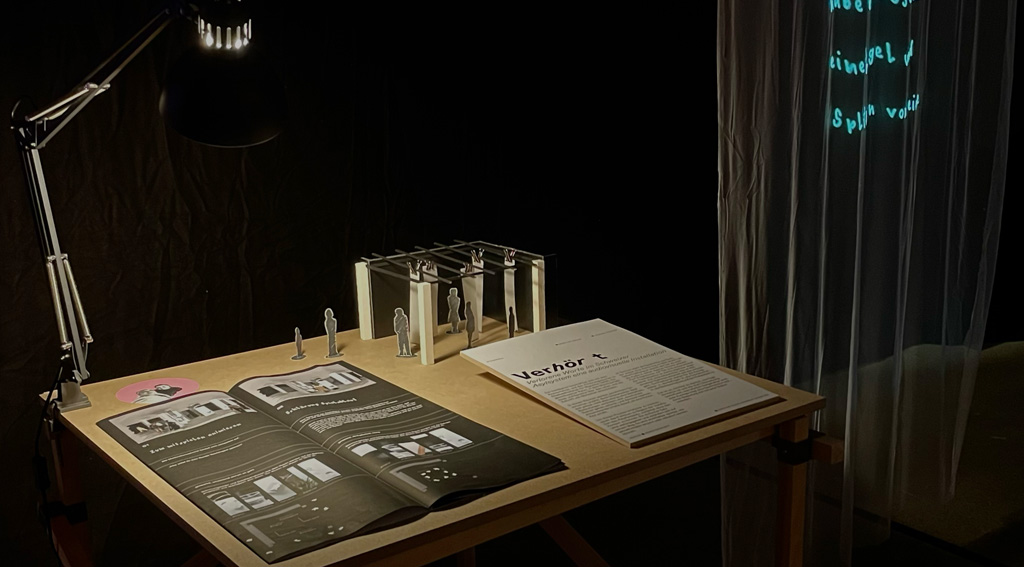 Dunkler Ausstellungsraum mit einem Tisch, darauf liegen Unterlagen und ein Arbeitsmodell – daneben hängt eine transparente Stoffbahn mit Projektion.